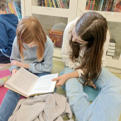 Zwei Schülerinnen lesen gemeinsam in einem Buch, im Hintergrund die Bücherregale der Bibliothek